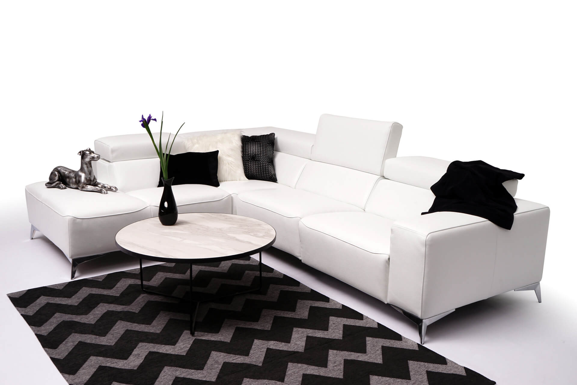 Narożna sofa Longo, obita białą sk&oacute;rą naturalną, wyposażona w funkcję relaksu i o wymiarach 229 x 297 cm, elegancko komponuje się z czarno-białym dywanem w zygzak i okrągłym marmurowym stolikiem kawowym. Całość uzupełniają elementy dekoracyjne, takie jak czarny wazon z kwiatami, srebrna rzeźba psa, poduszki i pluszowy pled.