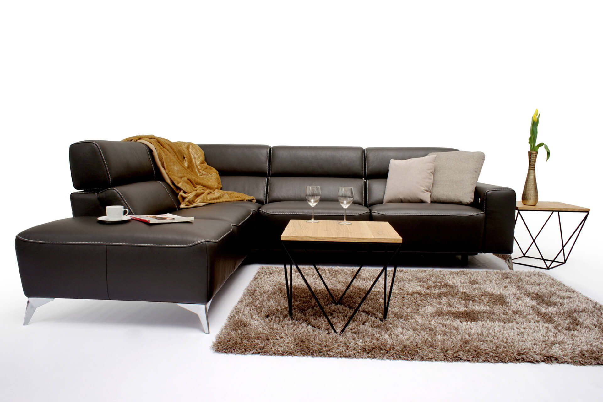 Nowoczesny salon prezentuje narożną sofę Infinity na wyższych nogach, wykonaną z naturalnej sk&oacute;ry. Czarna sofa jest wyposażona w beżowe i brązowe poduszki oraz musztardowy koc. Drewniany stolik kawowy mieści kieliszki do wina i czasopismo na pluszowym brązowym dywanie.